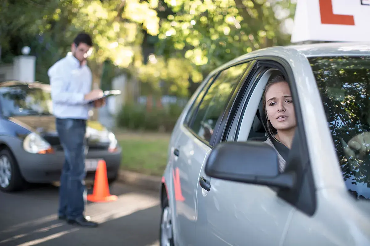 Joven nerviosa al volante, con un instructor anotando. ¿Se puede sacar el carnet de conducir sin autoescuela?