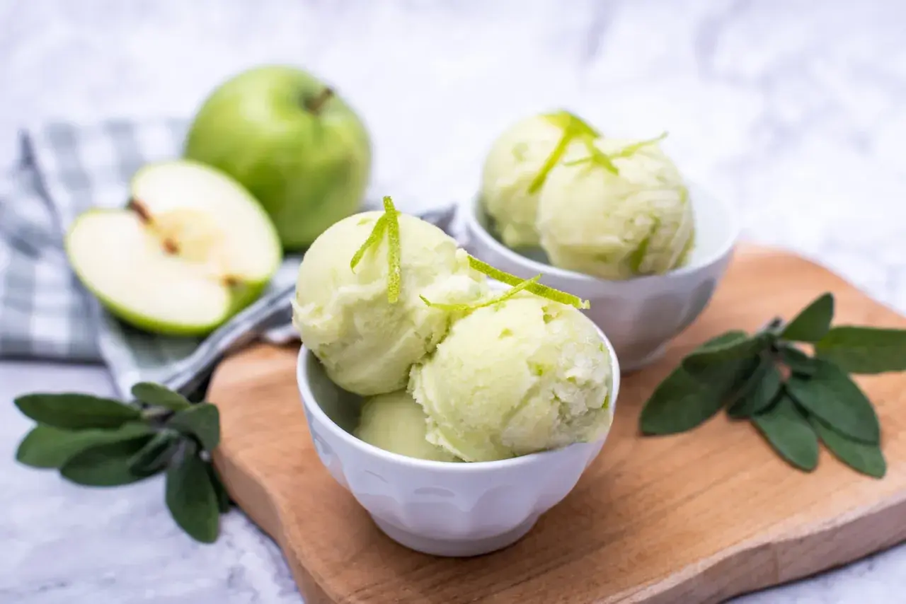 Orzeźwiający sorbet jabłkowy w miseczkach, udekorowany skórką z limonki. Obok całe i połówka zielonego jabłka.