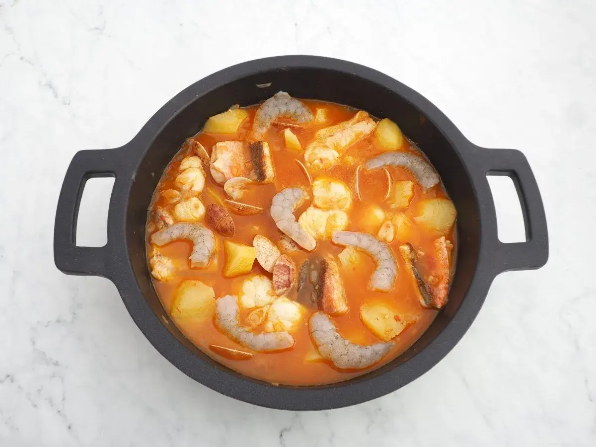 Un delicioso guiso marinero burbujea en una cazuela negra, con patatas, gambas, pescado y almejas en una salsa rojiza.