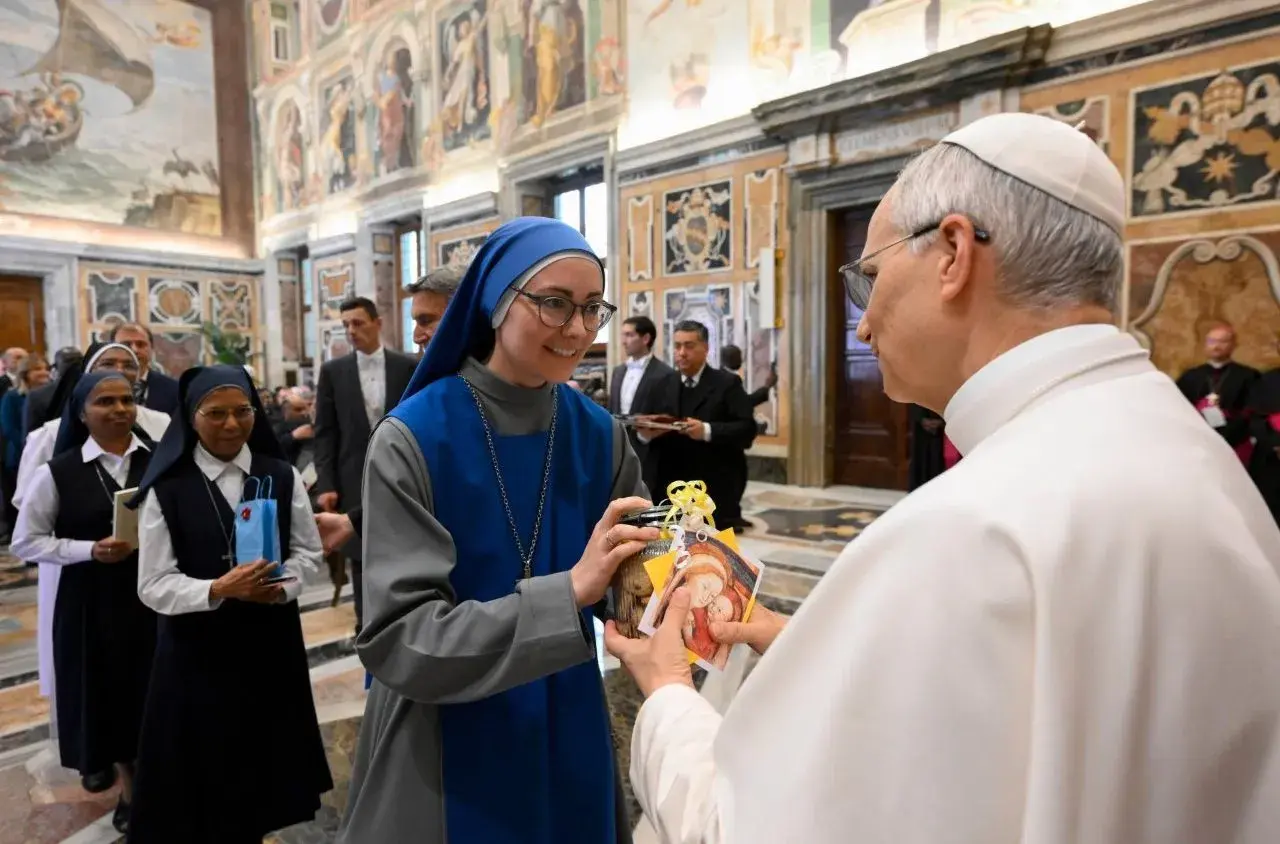 Eine Ordensfrau überreicht dem Papst ein Geschenk. Im Hintergrund stehen weitere Ordensschwestern und Männer in Anzügen.