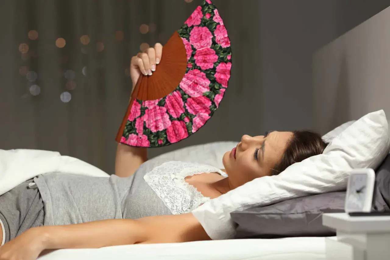 Donna a letto, insonne per il caldo, agita un ventaglio fiorato per combattere la sudorazione notturna.