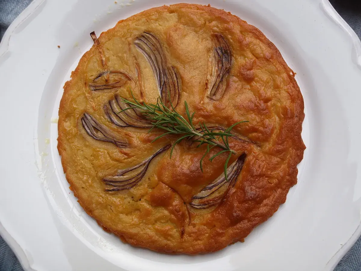 Herzhafte Kichererbsenmehl Rezepte: Goldbrauner Kuchen mit roten Zwiebeln und Rosmarin auf weißem Teller.