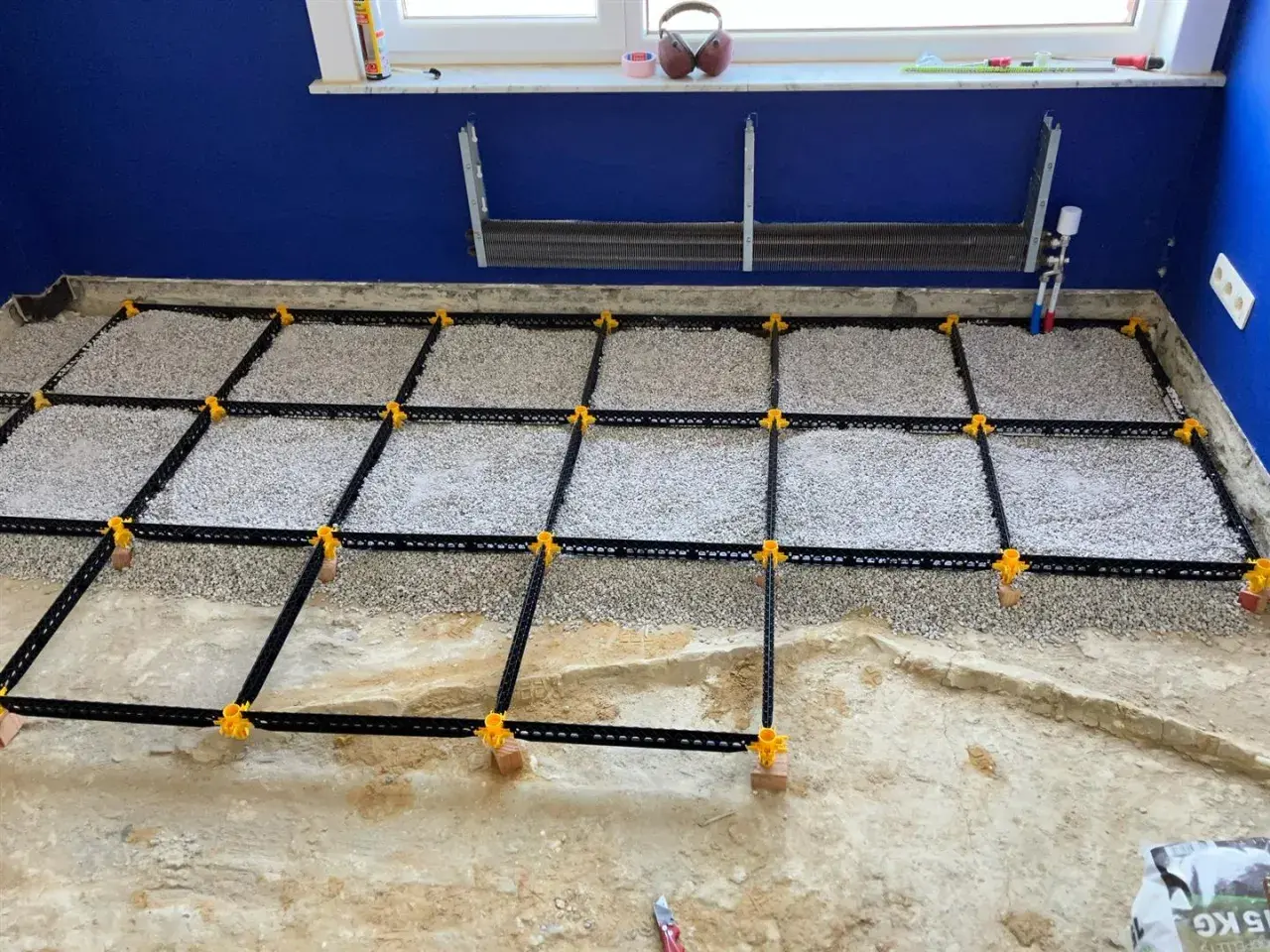 Vorbereitung für **OSB-Platten verlegen**: Ein Boden wird mit Schotter gefüllt und mit einem schwarzen Gitter und gelben Clips für den Trockenestrich vorbereitet.