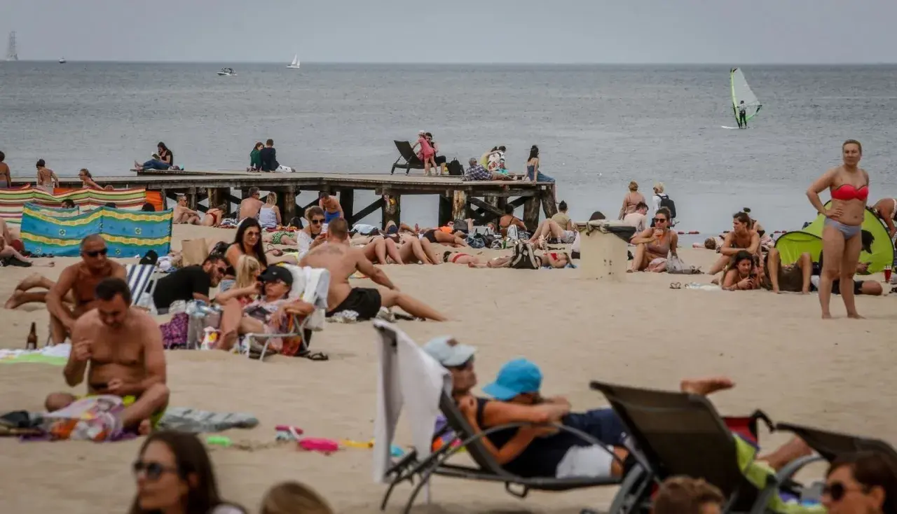 Tłum ludzi na plaży, opalanie się i relaks. Idealna pogoda na lipiec nad morzem, widać żaglówkę i wiatrak.