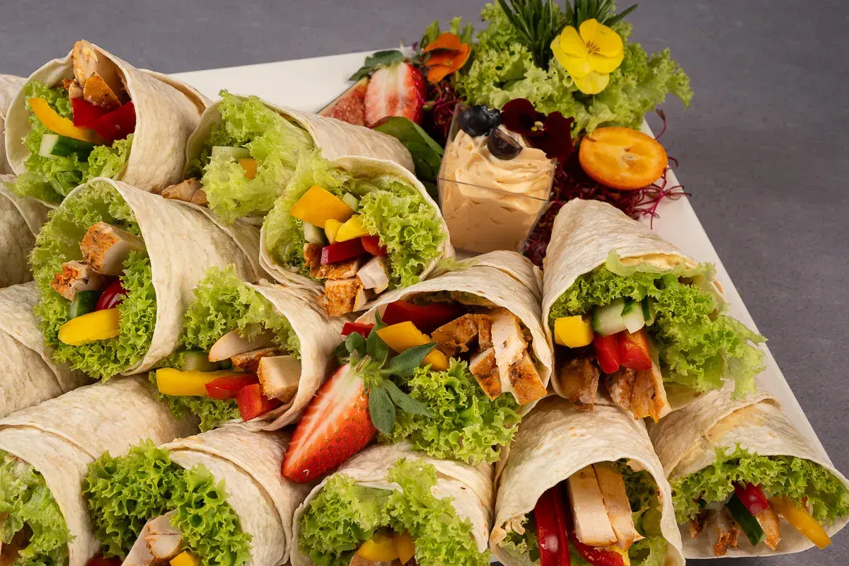 Mcwrap grillowany kurczak z warzywami i sałatą, ułożone na talerzu z dodatkami: dipem, owocami i ziołami.