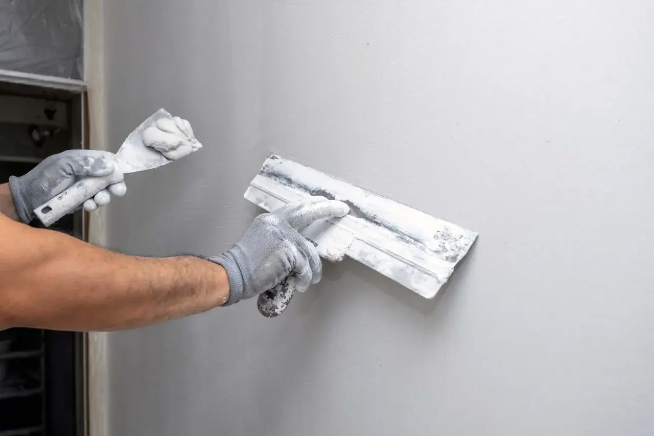 Mani guantate eseguono la rasatura di un muro con spatole cariche di stucco, preparando la superficie per la finitura.