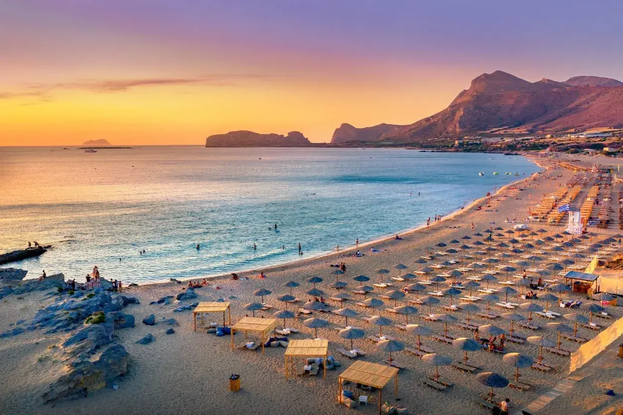 Ein langer Sandstrand mit vielen Liegestühlen und Sonnenschirmen. Das Meer ist ruhig, im Hintergrund Berge. Perfekt für die Frage: wo ist es im Mai warm Europa?
