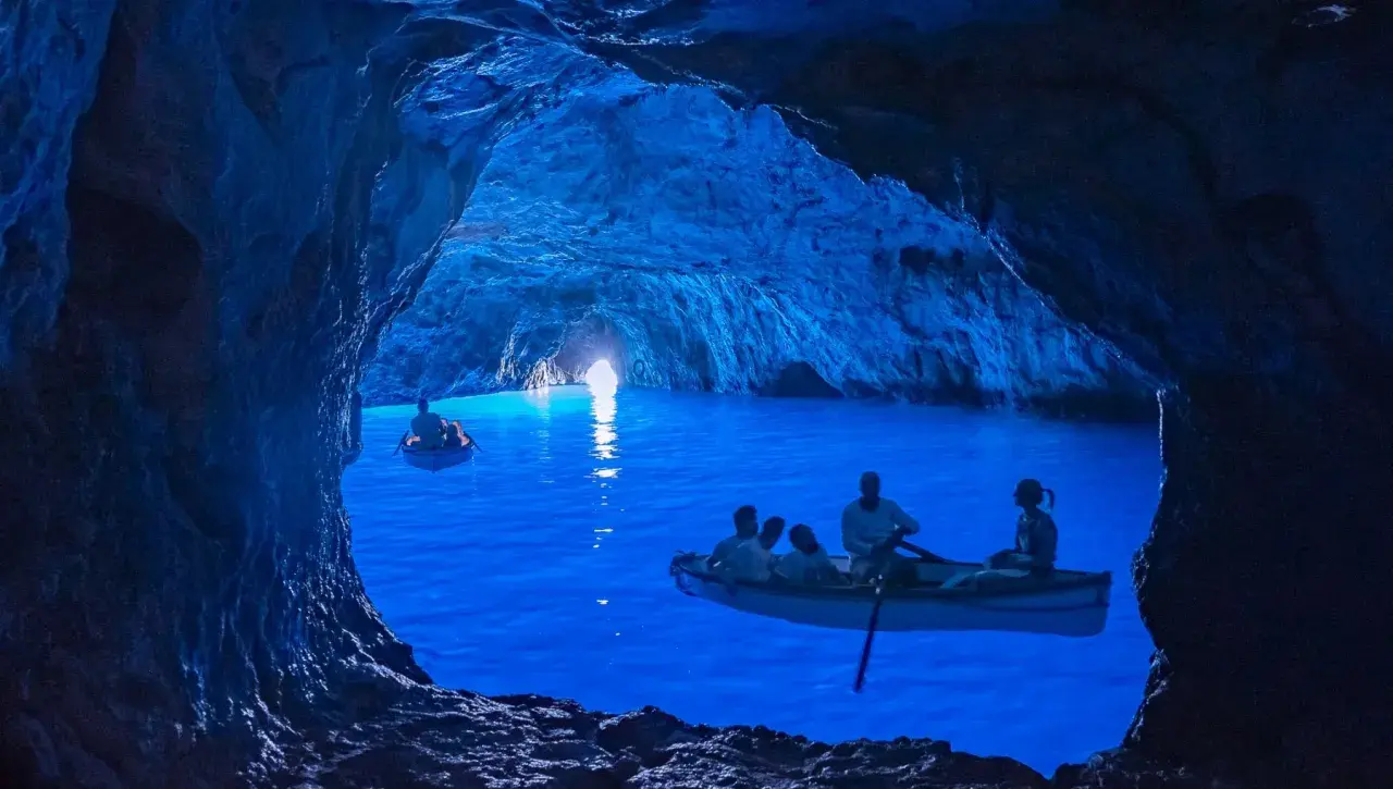Barche a remi navigano nelle magiche grotte blu, illuminate da una luce surreale che rende l'acqua di un blu intenso.