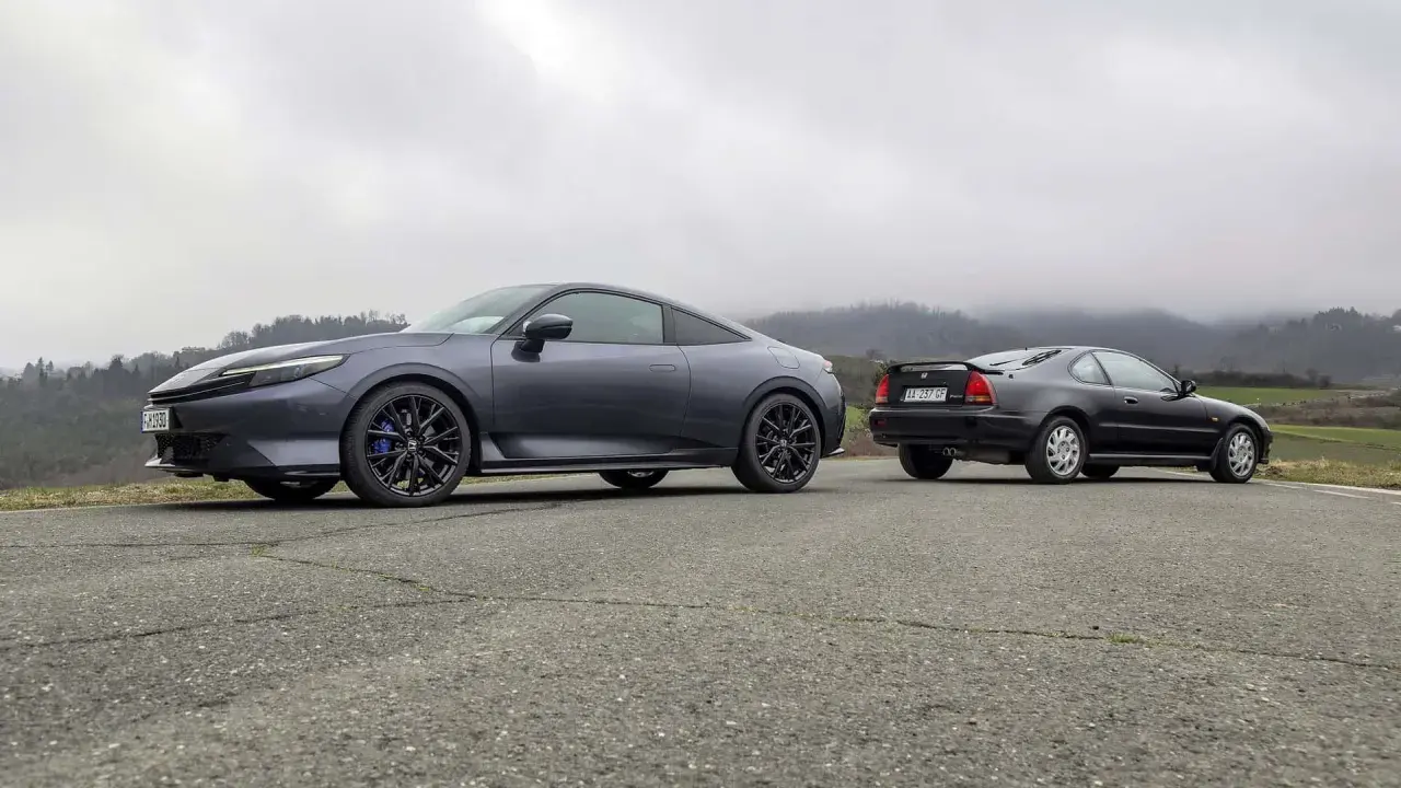 Due coupé Honda, una moderna e una classica, si incontrano su una strada di campagna. La nuova Honda Prelude 2026, grigio scuro, spicca accanto alla sua antenata.