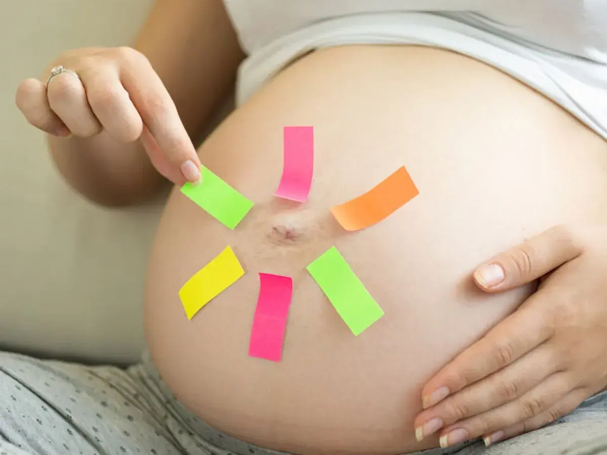 Une femme enceinte place des post-it colorés sur son ventre, peut-être pour choisir un prénom en P pour son bébé.