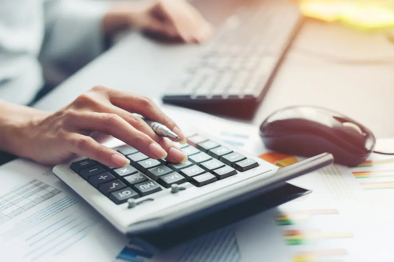 Dłoń z długopisem naciska klawisze kalkulatora, obok klawiatura i myszka. Analiza finansowa dla BNP Paribas Leasing.