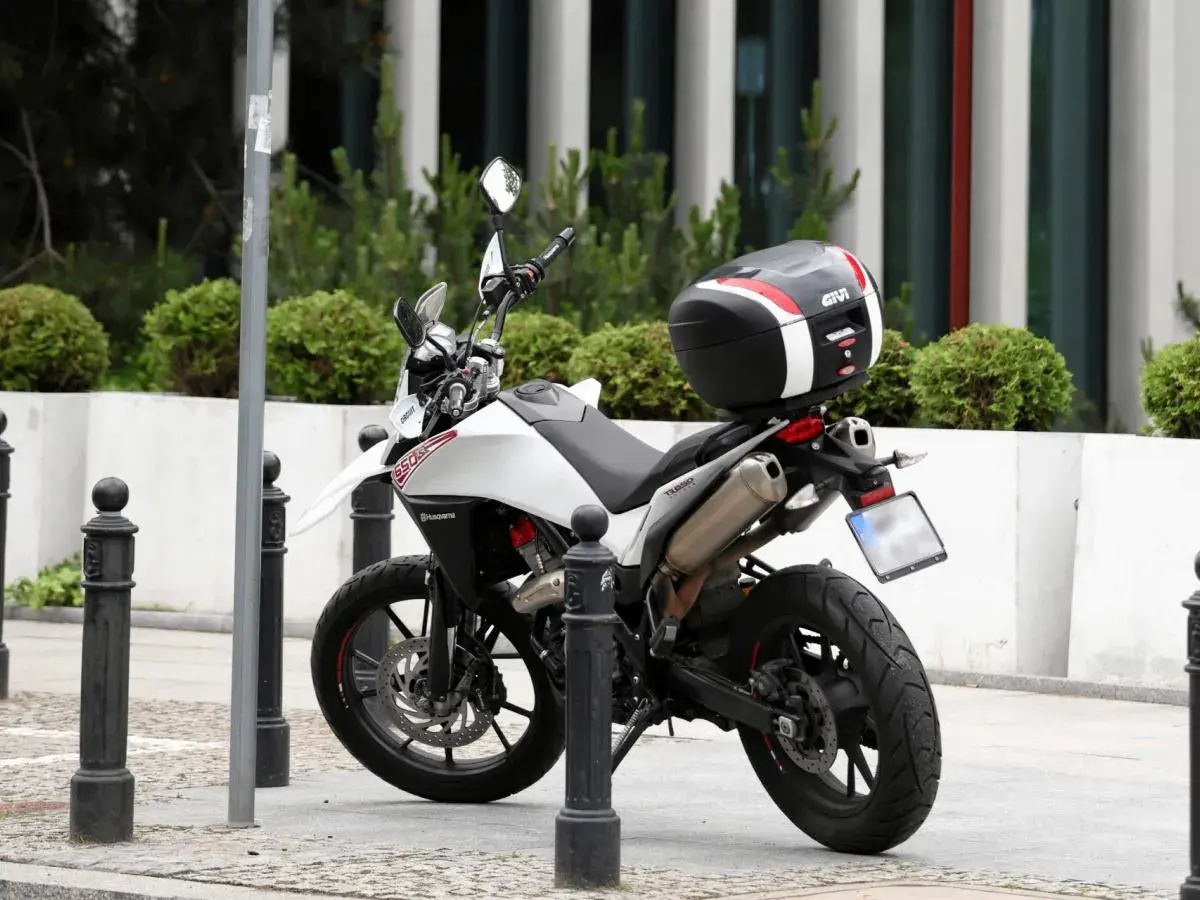 Biały motocykl z czarnym kufrem Givi zaparkowany na chodniku, obok słupków.