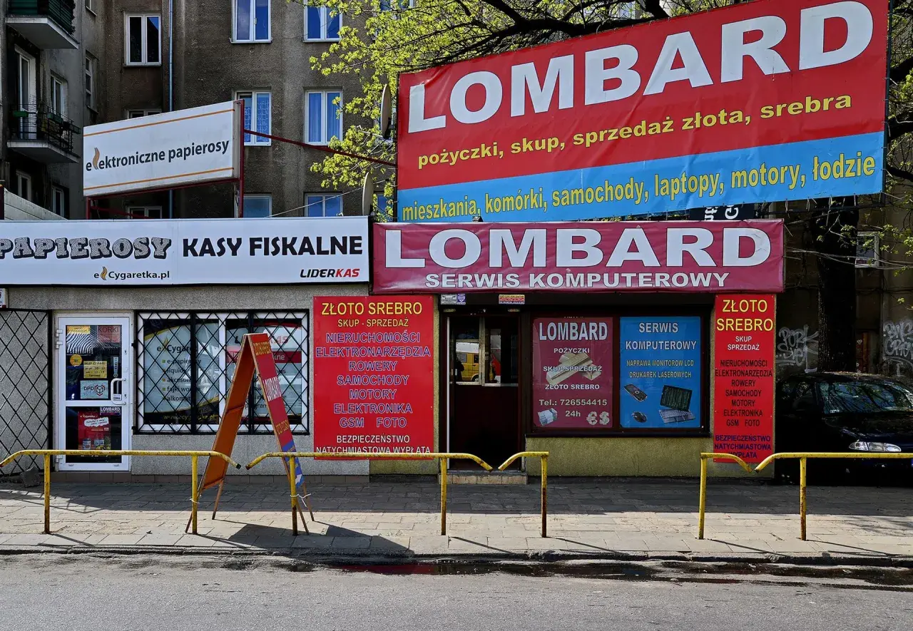 Szyldy lombardu i sklepów z elektroniką i papierosami na tle budynku mieszkalnego.