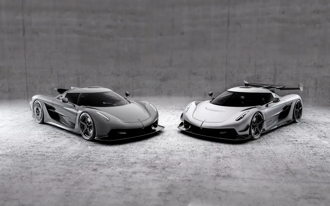 Dwa lśniące Koenigsegg Jesko, jeden szary, drugi srebrny, stoją obok siebie na betonowym tle.