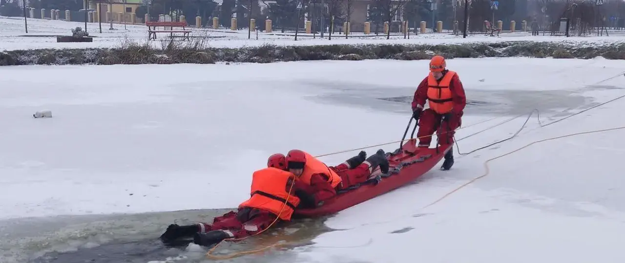 Ratownicy w akcji, wyciągają osobę z lodowatej wody za pomocą **sanki ratownicze**. Zimowa sceneria i profesjonalny sprzęt.