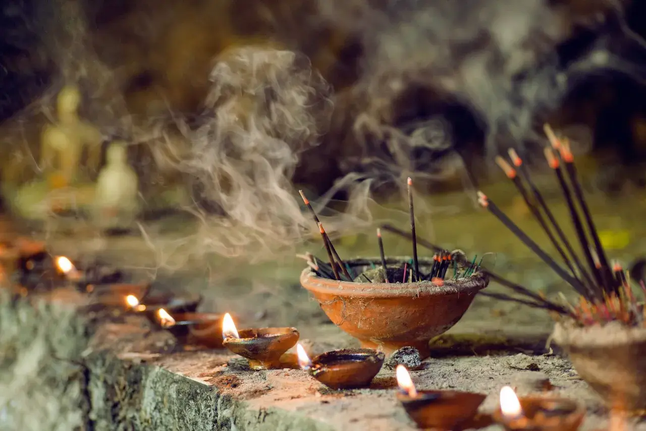 Humo de incienso llenando el aire, un recordatorio de que el incienso es malo para los pulmones. Velas encendidas y estatuillas borrosas en el fondo.