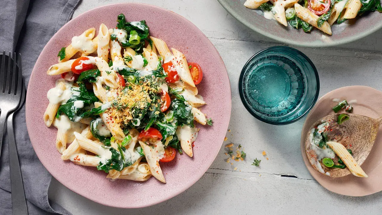 Penne mit Spinat und cremiger Sauce auf einem rosa Teller, garniert mit Kirschtomaten und Kräutern.