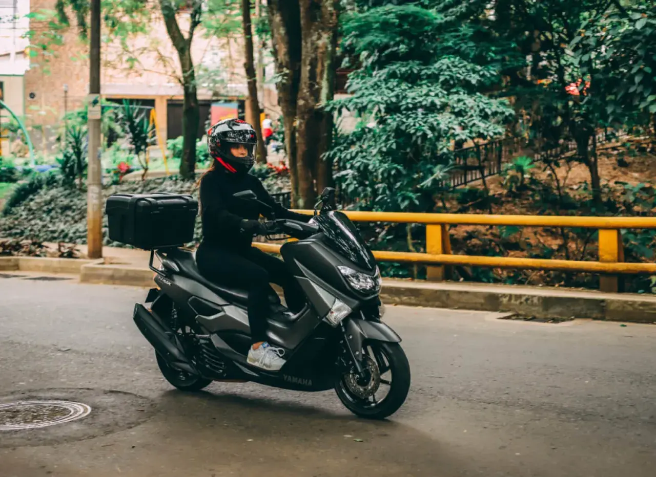 Mujer con casco y ropa negra conduce una moto Yamaha sin marchas, con caja trasera, por una calle arbolada.