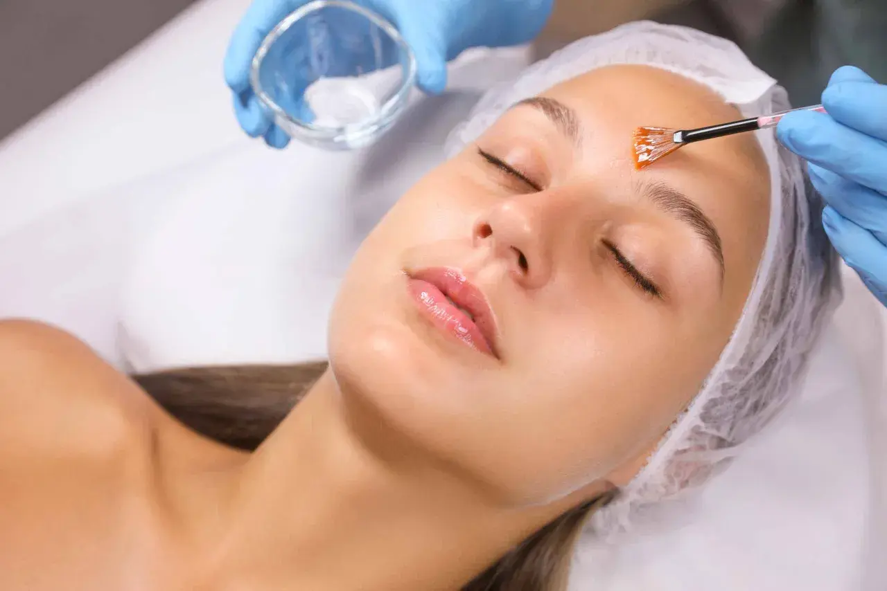 Manos enguantadas aplican un peeling facial con brocha a una mujer con los ojos cerrados.