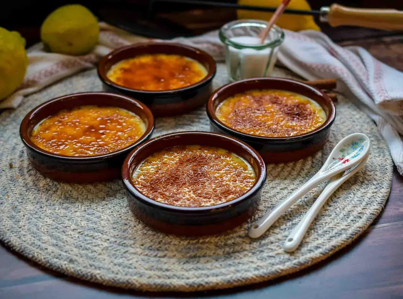 Vier Crema Catalana Schalen mit karamellisierter Zuckerkruste, bereit für das Crema Catalana Rezept. Zitronen und Löffel liegen daneben.