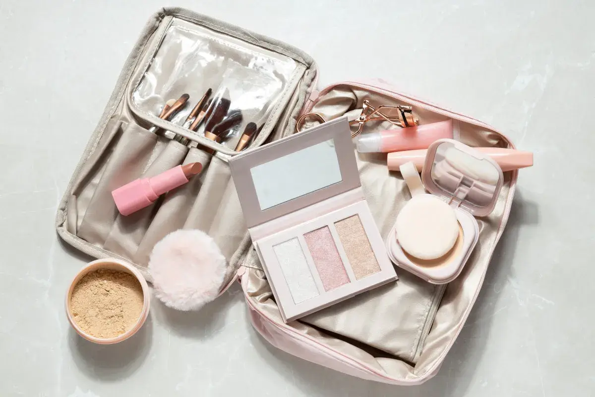 Une trousse de maquillage ouverte, prête pour un voyage en bagage cabine, révèle rouges à lèvres, pinceaux, palette et poudrier.