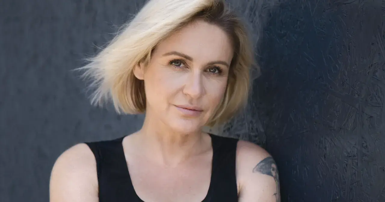 Małgorzata Ohme z blond włosami i tatuażem na ramieniu, na ciemnym tle.