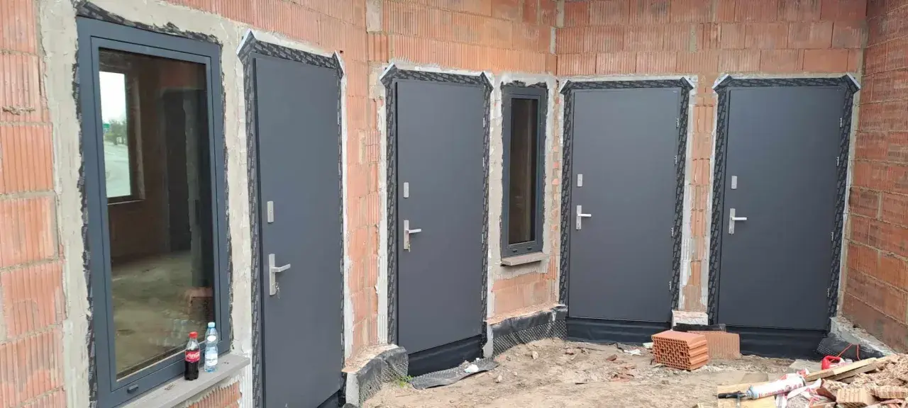 Nowe, szare drzwi zewnętrzne i okno w budowie. Widać ceglane ściany i materiały budowlane.