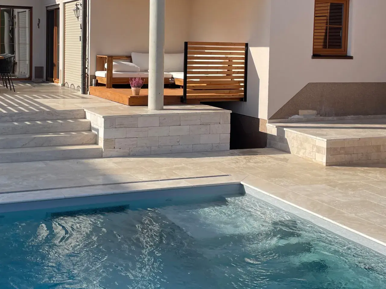 Piscina con bordo in pietra travertine. Gradini e pavimentazione in travertine creano un'elegante area relax con divani in legno.