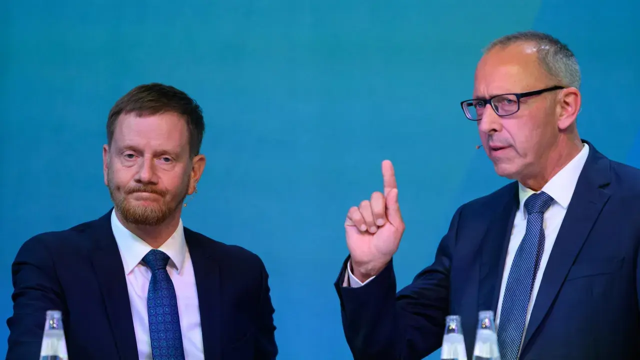 Zwei Männer im Anzug, einer mit Bart, der andere mit Brille und erhobenem Zeigefinger, diskutieren die Wahlprognose Sachsen 2026.