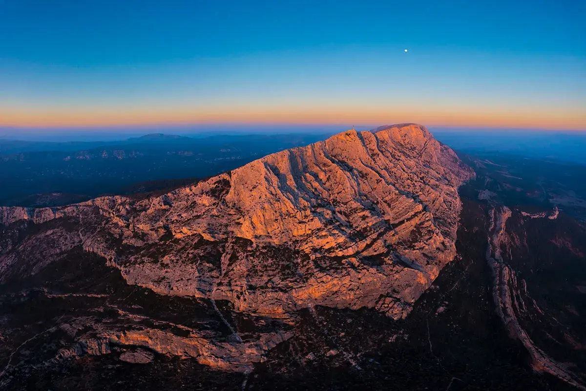 Le soleil couchant illumine la montagne Sainte-Victoire, offrant un spectacle grandiose pour les amateurs d'escalade. La lune veille sur ce paysage rocheux.