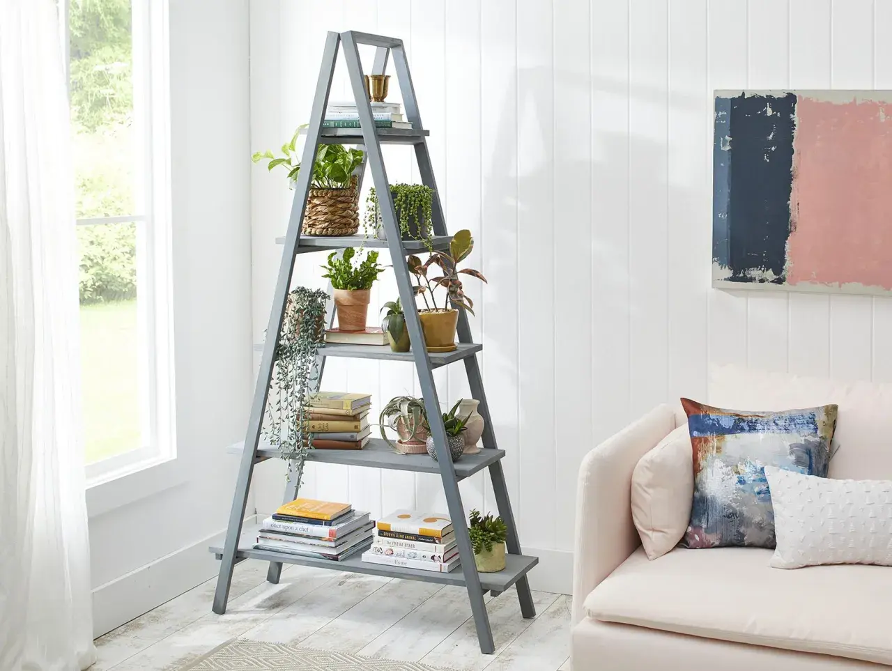 Perchero escalera gris con plantas y libros, junto a un sofá rosa.