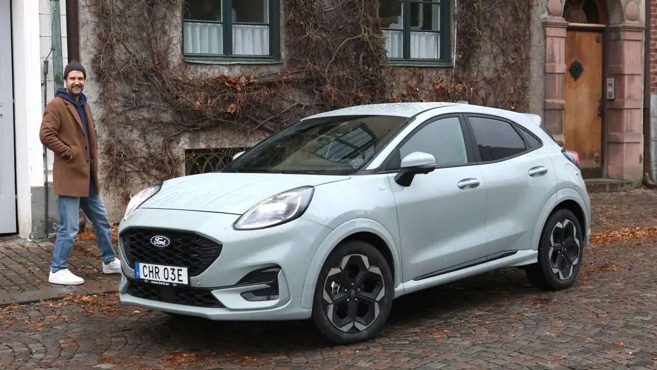 Man står bredvid Fords minsta modell, en ljusgrå Puma, framför ett gammalt hus med murgröna.