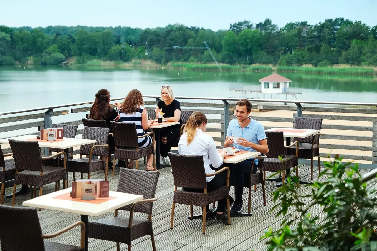 Gemütliches Restaurant am Wasser mit Gästen, die Kaffee und Getränke genießen. Im Hintergrund ein ruhiger See und grüne Bäume.