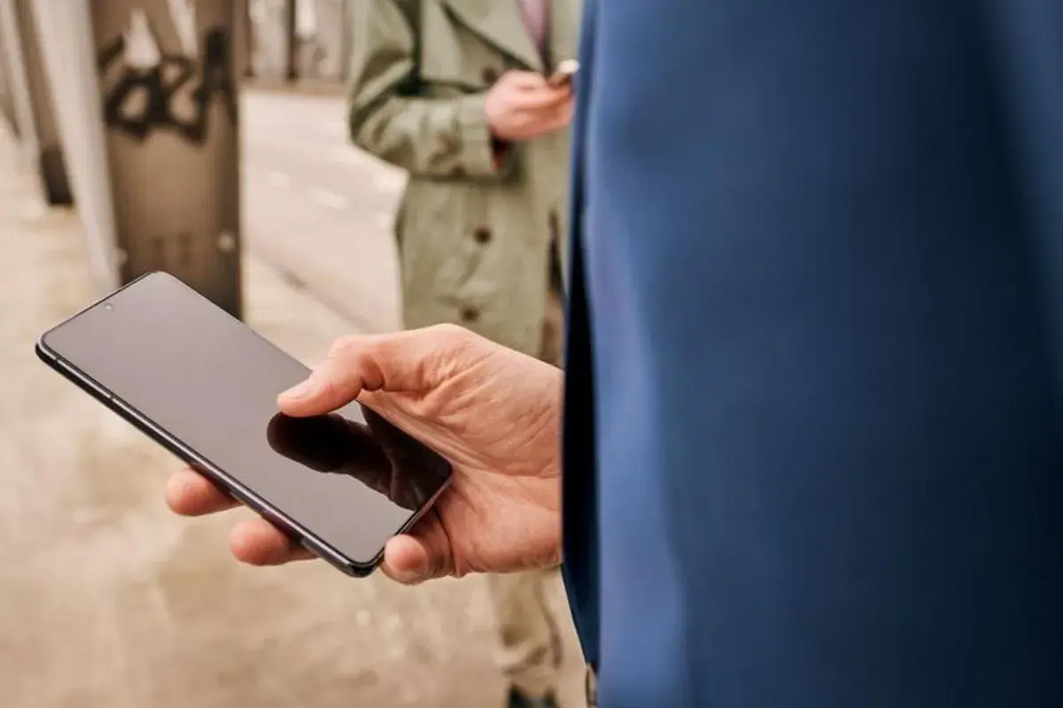 Mężczyzna trzyma smartfon, zastanawiając się, jak skonfigurować telefon. W tle druga osoba również korzysta z telefonu.