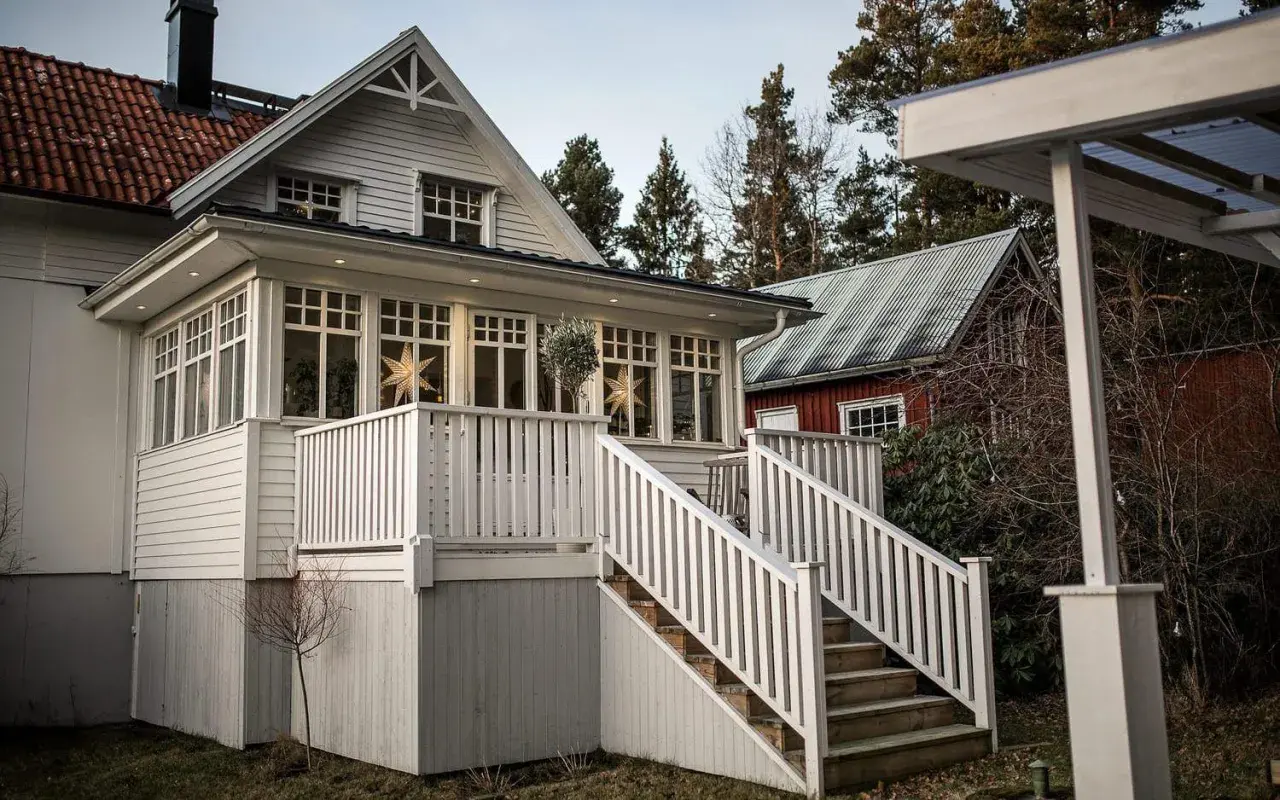 Vitt hus med glasveranda, stjärnor i fönstren och en röd stuga i bakgrunden. Glasveranda inspiration för ett mysigt hem.