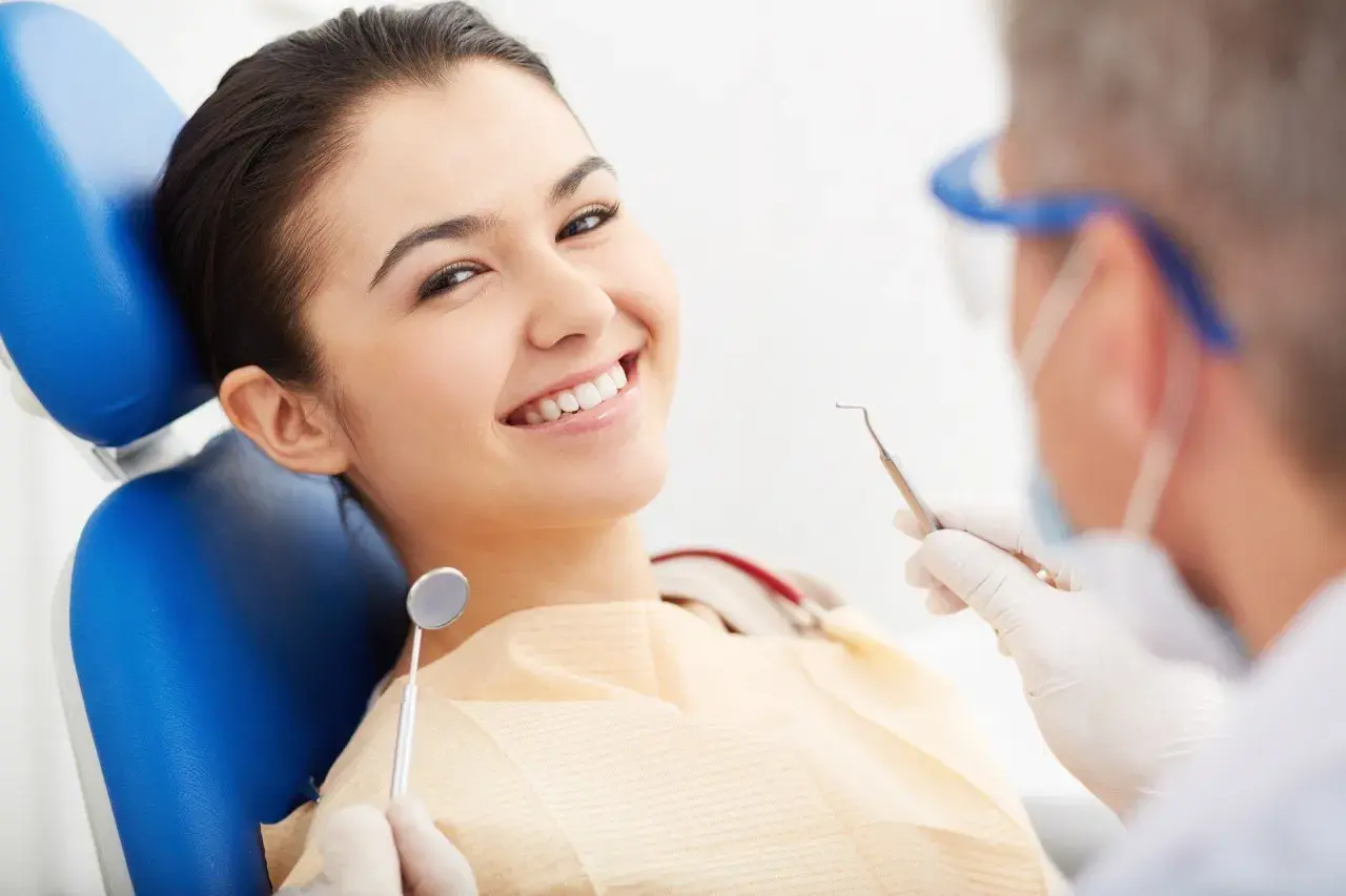 Uśmiechnięta pacjentka na fotelu dentystycznym, podczas gdy dentysta przygotowuje się do zabiegu z użyciem materiału glasjonomer.