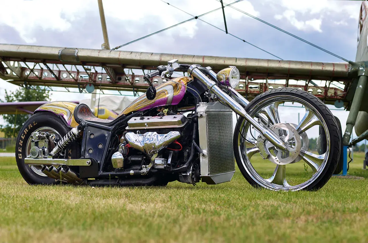 Ein glänzender Boss Hoss V8-Chopper mit lila-goldener Lackierung und einem riesigen V8-Motor steht auf einer Wiese vor einem Flugzeugwrack.