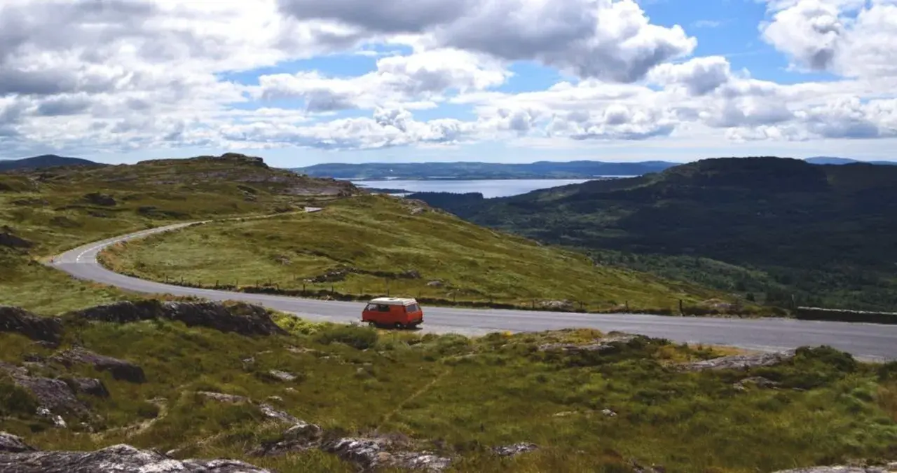 Eine malerische England Landschaft mit einer kurvigen Straße, einem roten Campervan und einem See in der Ferne unter bewölktem Himmel.