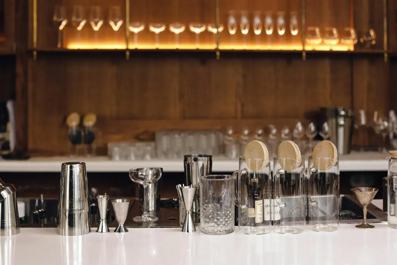 Na blacie baru stoją kieliszki, shakery i miarki. W tle widać rząd kieliszków do wina i drewniane półki. Każda miarka to jedna jednostka alkoholu.