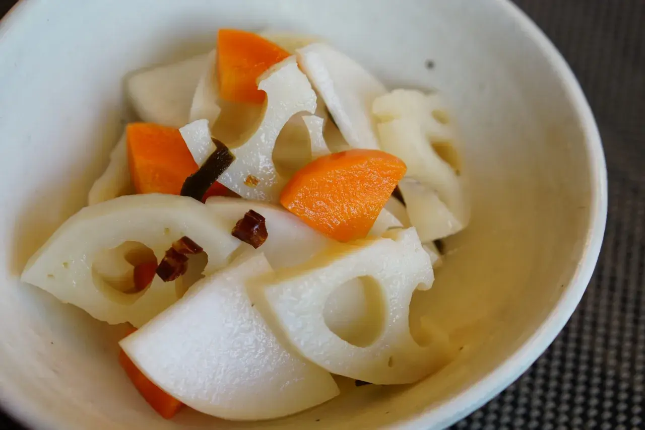 A close-up of a bowl filled with a colorful mix of Japanese root vegetables, including sliced lotus root with its distinctive holes, bright orange carrot pieces, and possibly daikon radish.