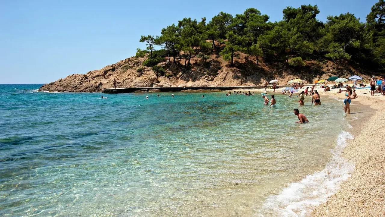 Słoneczna plaża na Thassos, z turkusową wodą i skalistym wybrzeżem. Ludzie kąpią się i odpoczywają, ciesząc się urokami wyspy. To idealne miejsce na wakacje, pełne atrakcji.