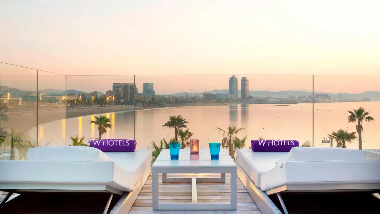 Dos tumbonas blancas con toallas moradas de W Hotels, una mesa con vasos y velas, y vistas al mar y la ciudad de Barcelona al atardecer.
