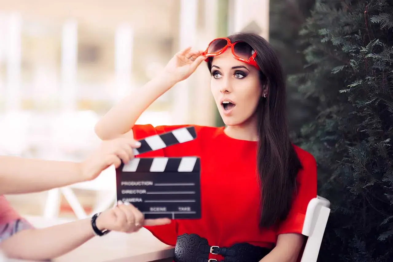 Aktorka w czerwonej sukience z okularami na głowie, zszokowana, gdy klaps filmowy zbliża się do jej twarzy. Stopniowanie high.