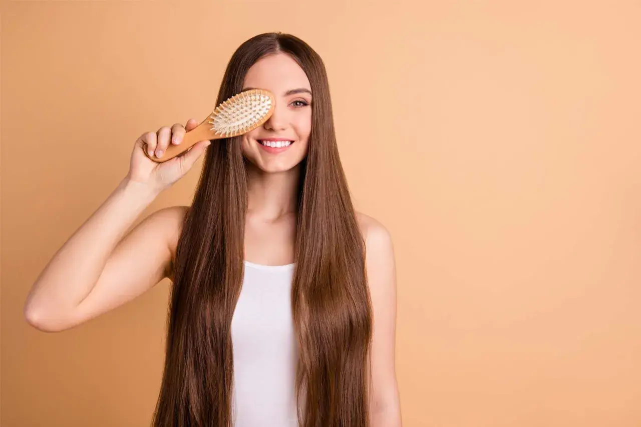 Mujer sonriente con pelo largo y liso, sosteniendo un cepillo de madera sobre su ojo. ¡Un truco para saber como desenredar un pelo muy enredado!