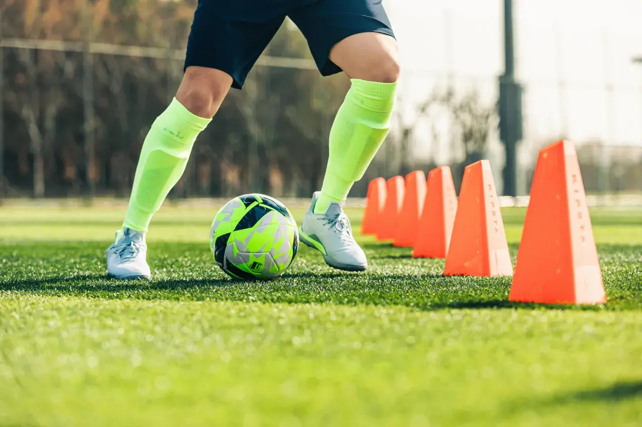 Piłkarz wykonuje ćwiczenia z piłką, mijając pomarańczowe pachołki na boisku.