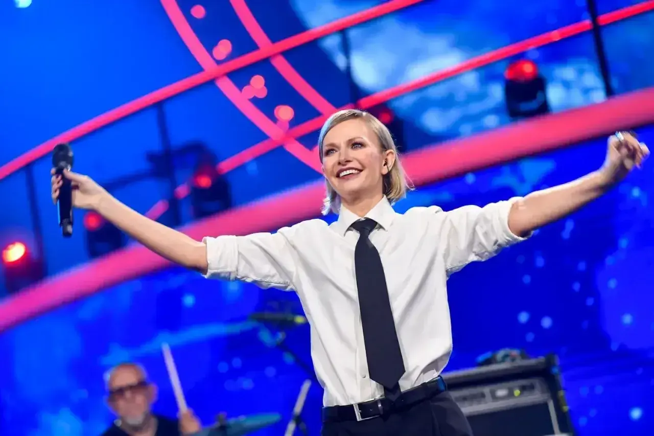 Kasia Stankiewicz na scenie, w białej koszuli i krawacie, śpiewa z mikrofonem. W tle niebieska scenografia z czerwonymi elementami, przypominająca orła cień.