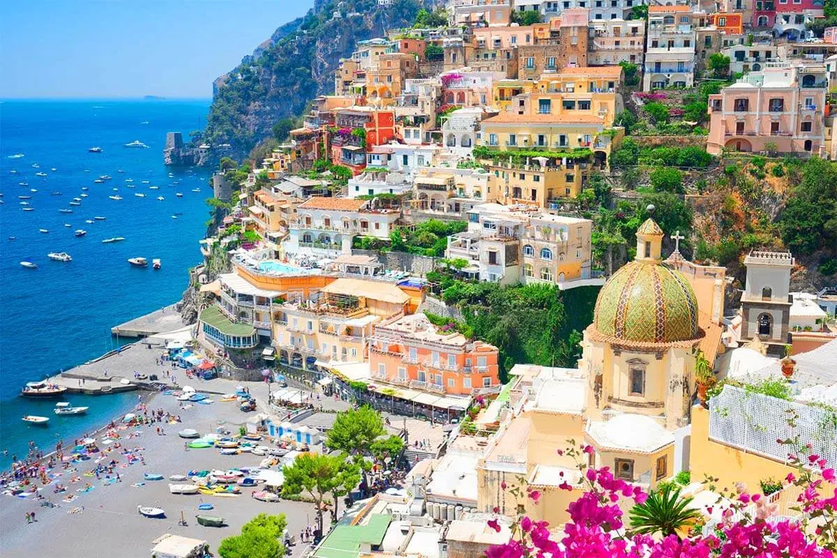 Colorful towns near Naples, Italy, cascade down a cliff to a busy beach and blue sea. A domed church stands out.