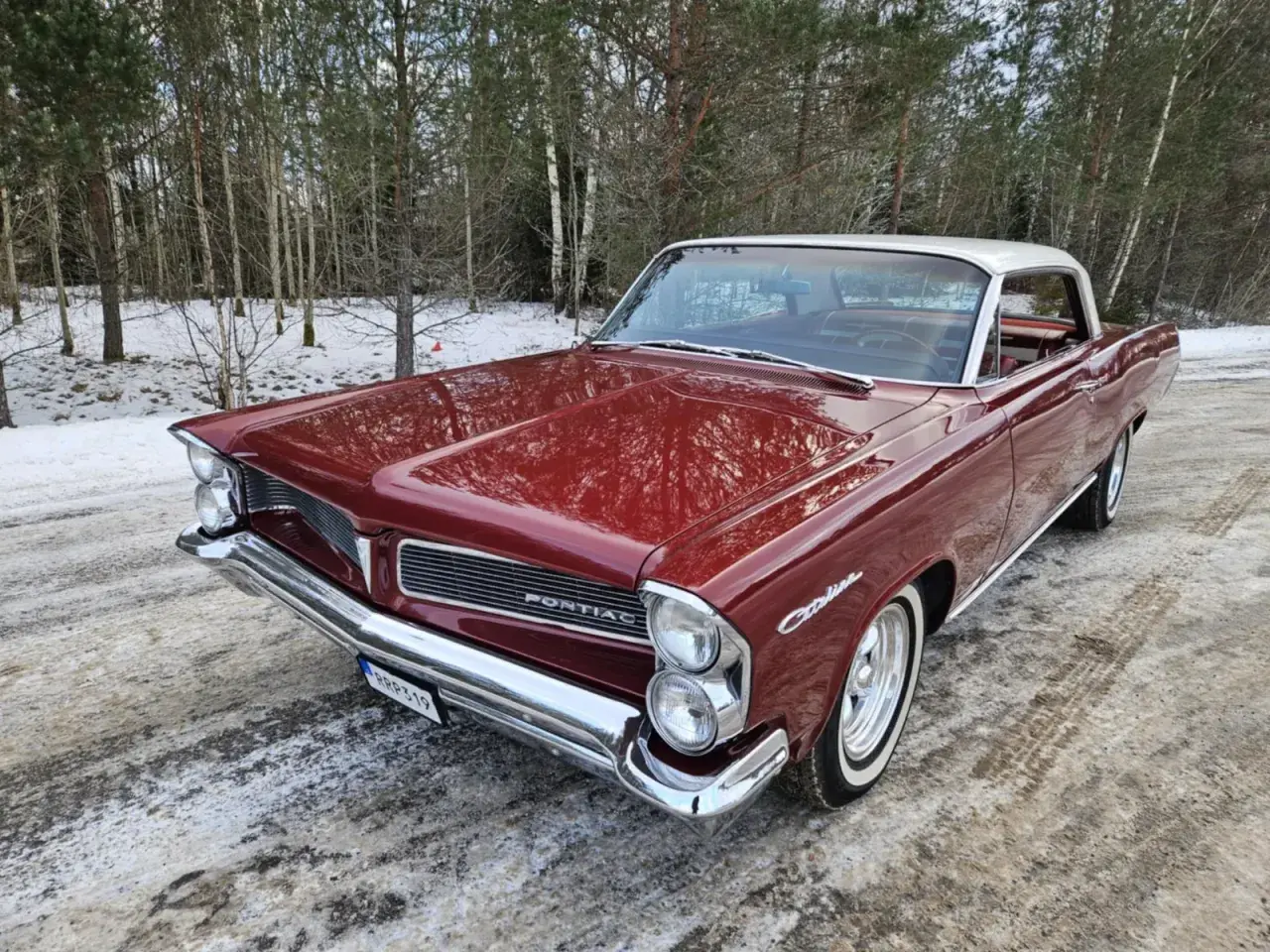 Röd Pontiac 60-tal, en klassisk bil med kromdetaljer, står parkerad i snöig skogsmiljö.