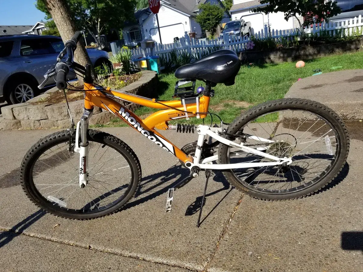 An orange Mongoose XR-75 mountain bike with a black seat and white suspension sits on a sidewalk.