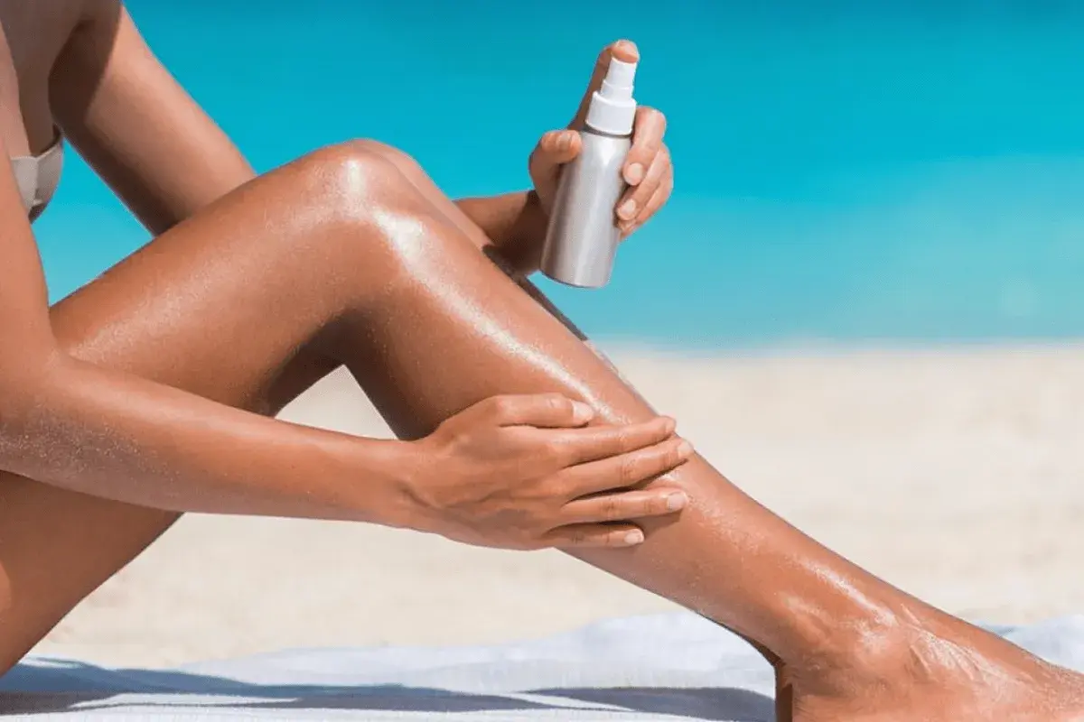 Donna applica spray abbronzante sulle gambe sulla spiaggia, per un'abbronzatura perfetta.